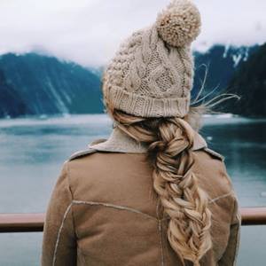 Femme aux cheveux blonds vêtue d’un manteau d’hiver et d'une tuque beige, debout au bord d’un lac en hiver.