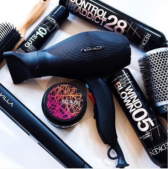 Séchoir à cheveux entouré de produits coiffants, de brosses et d’un fer plat, Credit photo: @redken